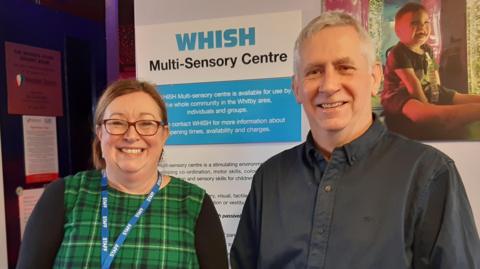 A woman wearing a green tartan dress and a blue staff lanyard stands smiling beside a man with grey hair wearing a dark shirt. Behind them is a picture of a giggling toddler in a multi-sensory room full of bright lights, beside a sign reading "WHISH multi-sensory centre".