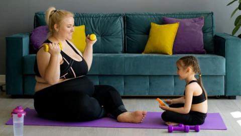 An obese pregnant mother holds dumbbells and sitting on a yoga mat. She is exercising with her little daughter. They are at home and facing each other on the yoga mat. They are smiling.