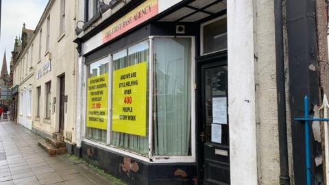 A store front in Dumfries with the sign First Base Agency in the window. It has black paint peeling at the bottom of a large window and crumbling window white window frames.
