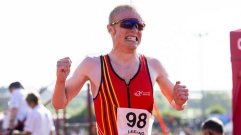 Corrin Leeming grits his teeth as he crosses a finish line. He holds both hands up by his sides in fists. He wears a red vest and wraparound sunglasses.