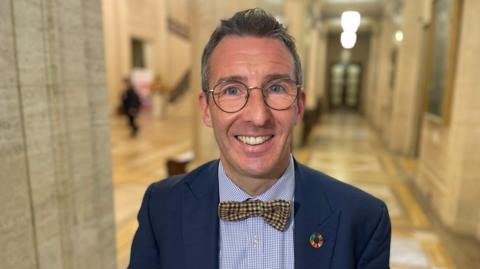 Andrew Muir is smiling. He wears glasses and has a navy jacket with a bow tie. He's wearing a colourful pin. He's in the hall in Stormont's parliament building. 