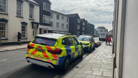 A row of buildings on a main road, two police cars parked next to the pavement,