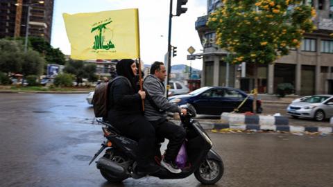 A side view of a woman carries a Hezbollah flag while riding pillion on the back of a motor scooter driven by a man on a street in Beirut