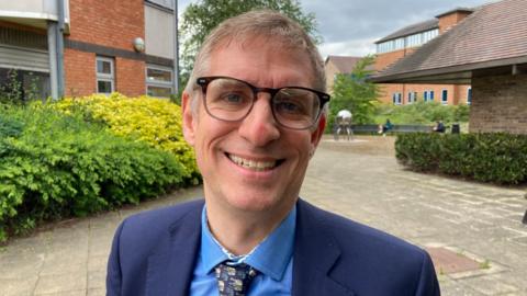 Councillor Matt Jenkins, wearing a blue suit jacket, blue shirt and patterned tie, is smiling at the camera.