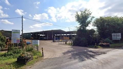 The entrance to a site showing a large open sided barn with construction vehicles, with signs saying Envar, its gates open