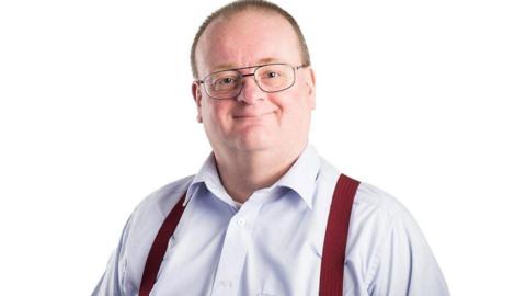 Phil Taylor, a man with dark, oval shaped glasses and short, light-brown hair. He is wearing a light shirt with dark red braces. He is looking directly at the camera and smiling.