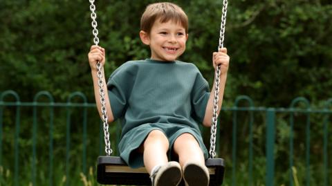 Elliot, a seven-year-old boy pictured on a swing outside. He has short brown hair and is wearing a green top and shorts. He is looking to the side and smiling. He has gaps in his teeth.