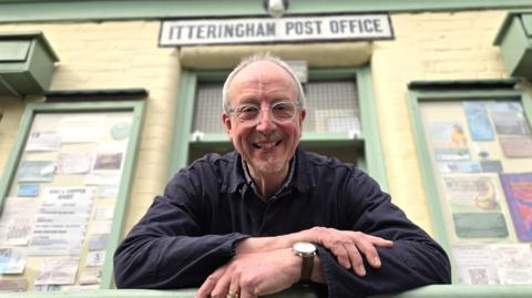 Mike Hemsley is leaning in front of a shop. He's wearing a dark blue shirt and transparently-rimmed glasses. Behind him is the 'Itteringham Post Office' sign.