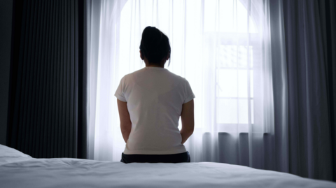 A woman is sitting on a bed. She's facing a window. Her hair is up. She's wearing a white top and dark trousers. Heavy drapes are parted. 
