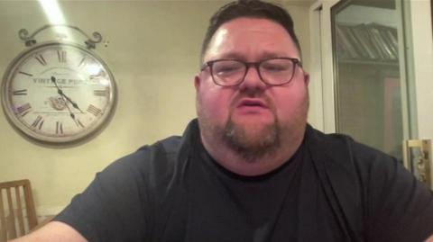 James Thomas, with a clock on the wall behind him, speaking about the market. He is wearing glasses and a black T-shirt and has short, dark hair and a beard.