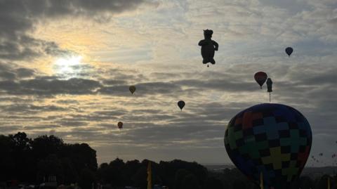 Eight hot air balloons, with one in the shape of a dragon and another as a screwdriver, silhouetted against the sky on a cloudy day.