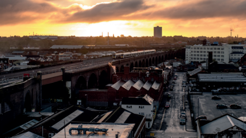 Bordesley Railway Viaduct can be seen as the sun sets in the distance. A train track runs on the left of the image with buildings and warehouses below to the right with a main street also visible.