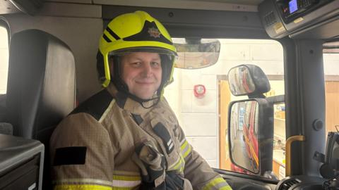 Sam Heslop dressed in firefighter uniform sat in the passenger seat of a fire engine.