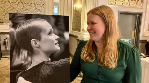 Mum Vicky Robayna, who has mid-length blonde hair, looks at a large black and white photograph of her teen daughter Liz Hatton. 