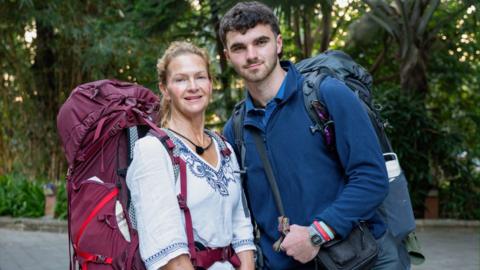 Caroline and Tom are both wearing large backpacks. They are standing together in front of shrubbery.