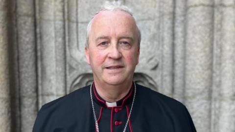 A man in black and red clerical garb stands in front of the pillars of a church. A cross is being worn around his neck. 