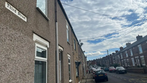 Terraced housing with the road sign saying Third Street. Cars are parked on both sides of the street.
