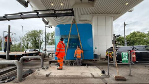A blue toll booth being held up by a small crane and sat on a trailer. To the right of it is a black car and to the left is a crane. There is a white building above it. The sky is grey and cloudy. 
