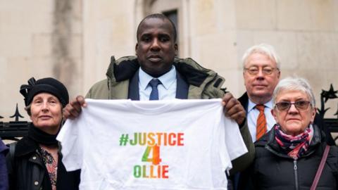 Oliver Campbell wearing a suit, standing outside the Royal Courts of Justice in London. He is flanked on both sides by campaigners for his case. He is holding a white t-shirt emblazoned with #justice4Ollie