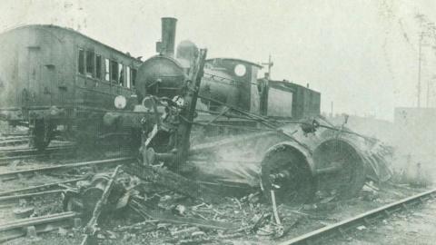 The Witham railway crash of 1905. A black-and-white image shows the remains of a railway carriage, with wheels and iron framework surrounded by broken pieces of wood, sitting on railway tracks. Smoke is rising from the wreckage, and there is a locomotive engine in the background.