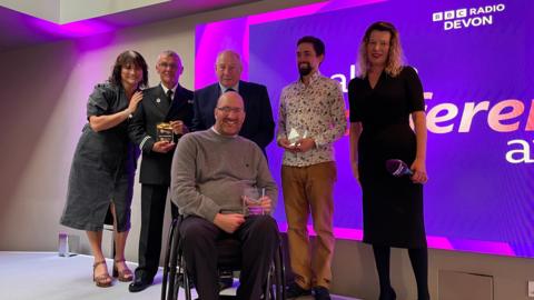 Six people are in a row with one person slightly in front of them, some are holding awards which appear to be made of glass, and host Angela Kalwaites is one of the people who is holding a microphone 