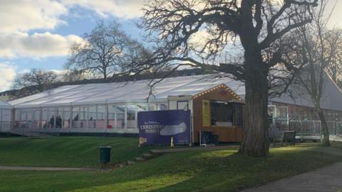 The ice rink in Imperial Gardens, Cheltenham. It is a large white marquee on a grassy field with festoon lighting and an advertisement sign for the Christmas Market. 