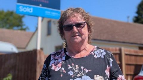 Viv Matthews poses in front of her home. She is wearing a floral dress and has short, curly hair and dark sunglasses. A 'sold' house sign can be seen behind her