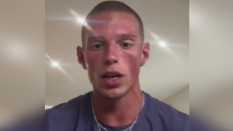 Finn speaking into a camera during the interview. He has very closely shaved hair and is wearing a purple shirt with a silver chain.