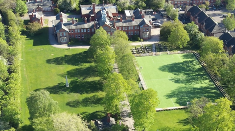 An aerial view of an ornate building with a large field in front of it as well as a playing field and trees. There are rows of houses to the left hand side of the image.