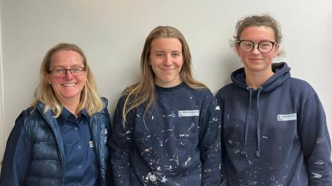 Jane, Faith and Antonia stand side by side against a plain light-coloured wall. They are wearing dark blue work clothing with visible paint splatters. Faith and Antonia are in hooded sweatshirts with company logos that read “Bagnalls,” while Jane is wearing a padded vest over a collared shirt.