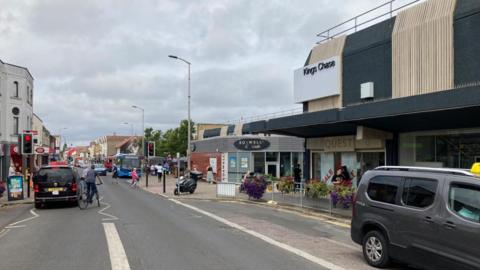 The photo shows Kingswood High Street under cloudy skies. Kings Chase shopping centre can be seen on the right, wit Boswell Cafe in the distance. Lots of people can be seen walking on the pavements, while a small number of cars and a cyclist are stopped at a red light.
