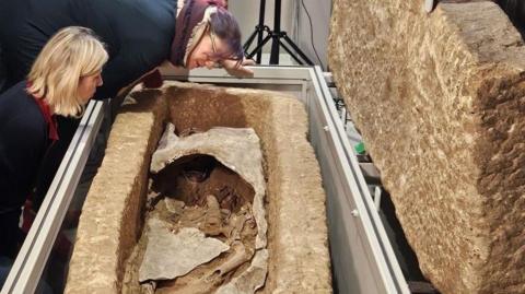 Two women inspect a fragmentarily preserved gypsum burial in a stone coffin