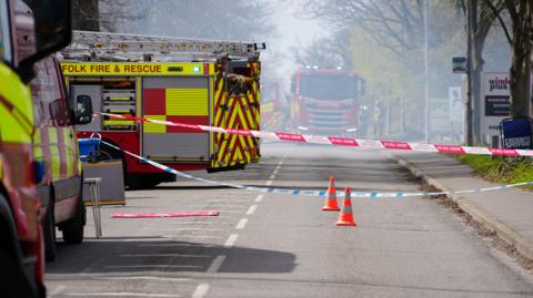 Red-and-white and blue-and-white cordon tape runs across a road. Two orange traffic cones are in the middle of the road, with fire engines parked to the left and in the background. The air appears misty with smoke.