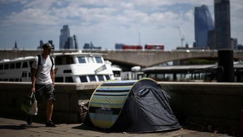 A small striped Trespass tent set up on a paved embankment along the River Thames, as a young man carrying shopping bags walks by, with riverboats, a bridge featuring red double-decker buses, and London skyscrapers visible behind.