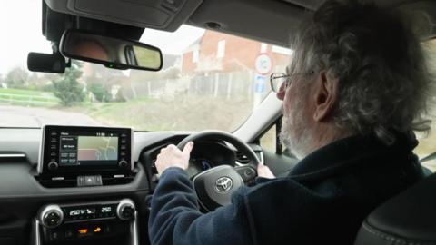 Photograph of Turner driving, taken over his shoulder. You can see the back of Turner's head - he has short, curly grey hair and a beard. He is wearing a pair of thin-framed glasses, and a dark blue fleece. The interior of his car is black, with a silver Toyota logo on the wheel. Outside the windscreen, you can see Turner is driving down a residential street. 