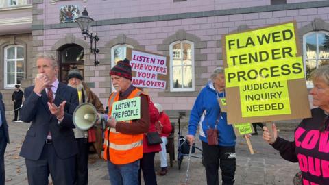 Protestors from Love Our Lido gather outside the States Assembly. One man is in a suite and speaking into a loudspeaker. Other protesters are holding placards that say "flawed tender process" and "listen to the voters".