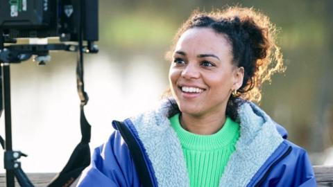 Cat White, a filmmaker, sitting down looking into a camera monitor. She is wearing a bright green jumper with a blue fleece-lined coat over the top. She is smiling but not looking at the camera.