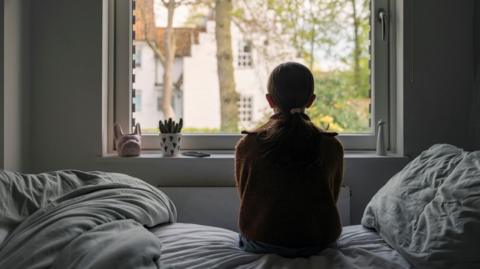 A girl is sitting on a bed with her back to the camera looking out of a window. She is wearing a brown cardigan and has brown hair tied back.