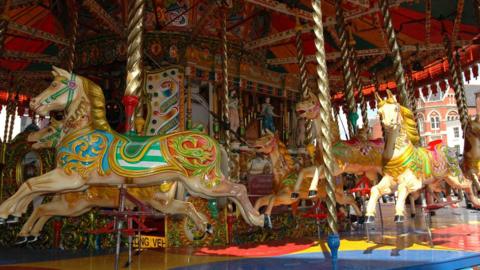 A carousel can be seen with several yellow horses with colourful saddles on them. Swirly gold posts can also be seen.