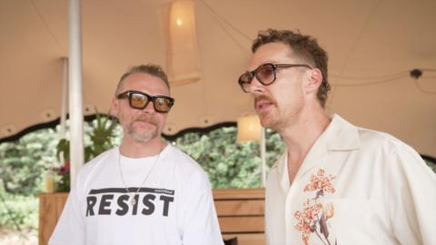 Simon Pegg and Benedict Cumberbatch stand side by side under a tent canopy. Pegg is smiling and laughing as he looks towards Cumberbatch. They are both wearing sunglasses. Pegg has on a white T-shirt which says 'Resist' in upper case, black letters. Cumberbatch has on a cream, short-sleeved shirt with a floral design on one side.