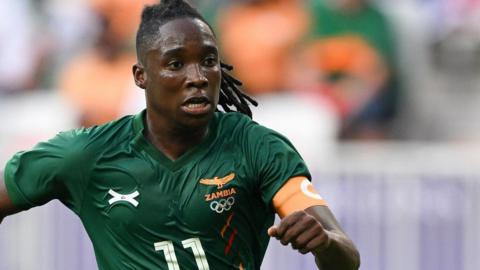 Barbra Banda, wearing a green Zambia shirt with the number 11 on it and a yellow captain's armband, runs forward with a purposeful look on her face during a football match at the Paris 2024 Olympics