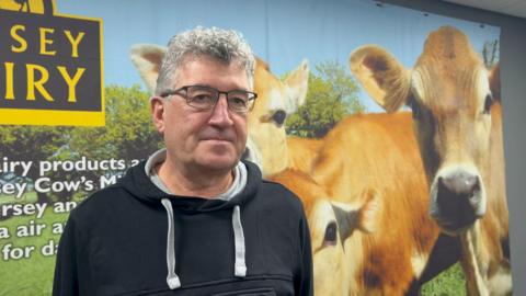 Eamon Fenlon - a man with black rectangle glasses wearing a black hoodie standing in front of a poster of Jersey Dairy with three cows on it.