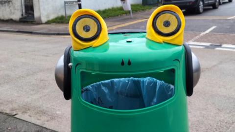 A close-up of a green bin with two yellow eyes on the top and a set of headphones. The opening of the bin is designed to look like a mouth, with a nose drawn above it.