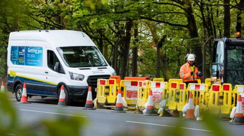 White van with "Getting gas to you" written on its side. There are cones and  tractor and a man with PPE working next to a road with trees in the background.