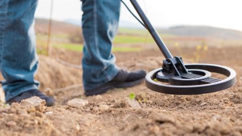 A man's legs are in shot as he holds a metal detector on a soil ground. He is wearing blue jeans and black trainers.