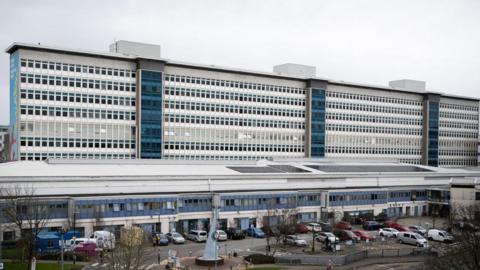 The outside of a large hospital, which is tall, blue and white with many windows, and car parked in front of it.