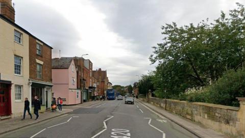 Road with buildings along the left and a stone wall along the right.  Buses and cars are in the distance and people are walking on the pavement.