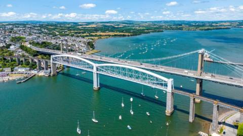 The Tamar Bridge shown with blue water underneath and boats dotted on the water. There are cars driving along the bridge. 