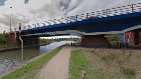 A view of the blue canal bridge from a footpath below it, on an overcast day.
