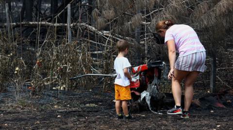 A woman and a toddler stand by charred ground and burned plants.